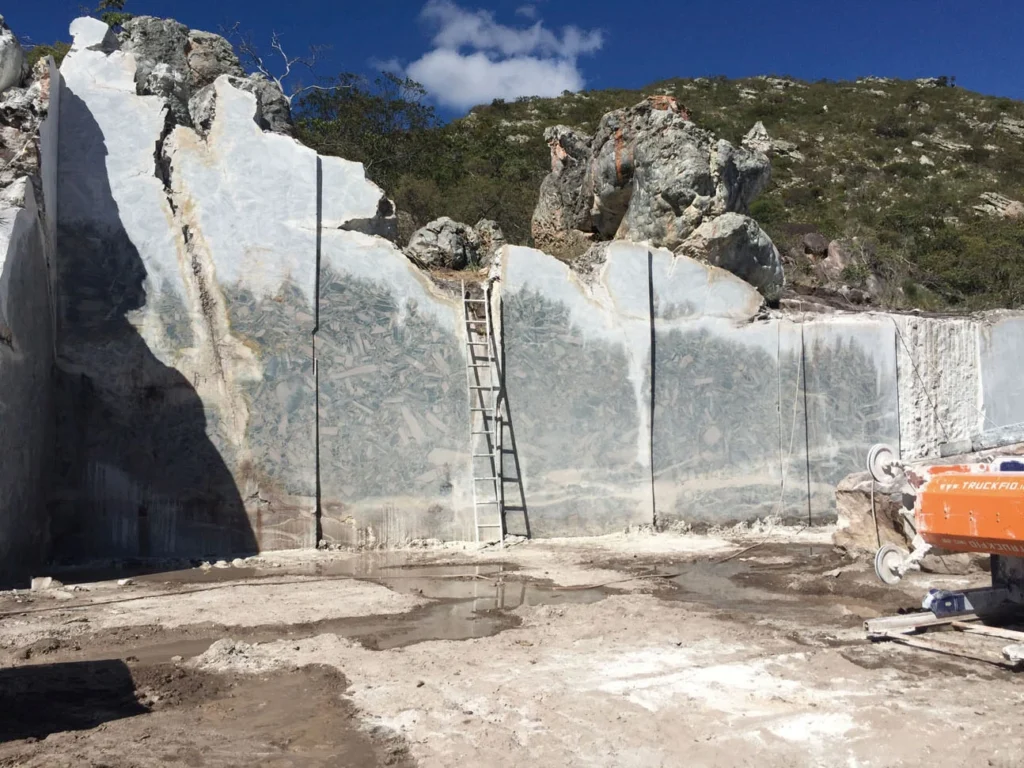 Open-pit marble quarry – Massive white marble cliffs with natural textures, a worker on-site, set against a clear sky and green hills.
